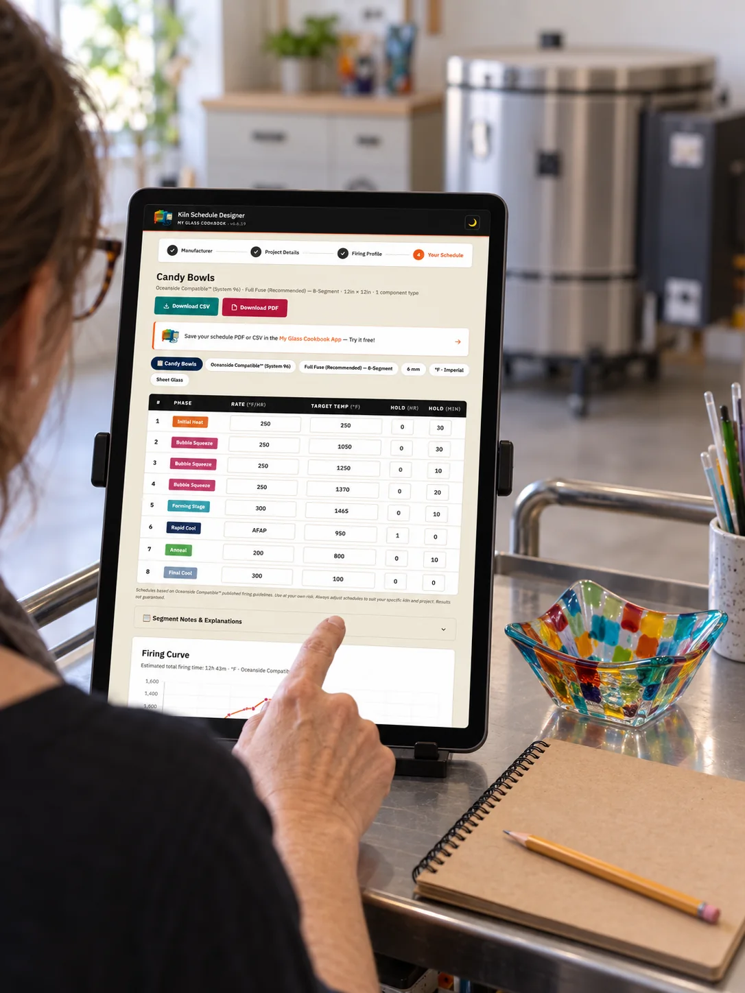 Artist viewing a firing schedule on a studio tablet beside a colorful glass bowl and notebook, with a kiln behind